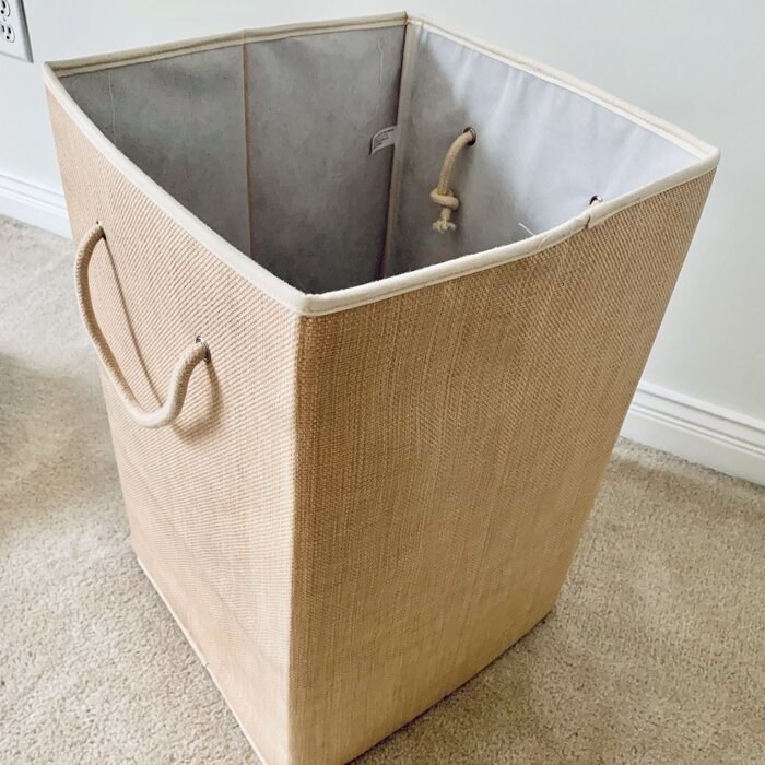 A light natural tan wicker laundry hamper with rope handles photographed on the carpet of a reviewer&#x27;s home