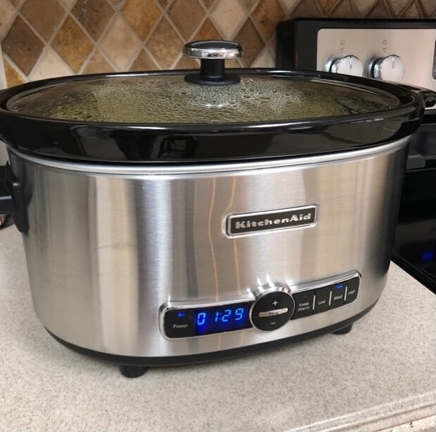A metallic silver slow cooker with a black ceramic pot turned on showing condensation under the see-through glass lid