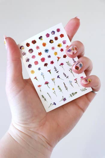 A person's hand displaying a sheet of 60 tiny flower tattoos for nails 