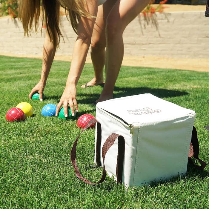 a person bending down in grass to pick up red, green, yellow, and blue bocce balls next to a white zippered square case to hold the balls in