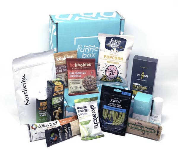 A blue box that says &quot;The RunnerBox&quot; with snacks and running accessories in front of it including a white package that says &quot;Lesser Evil Organic Popcorn&quot; and a bottle that says &quot;Tiger Balm Active Muscle Spray&quot; 