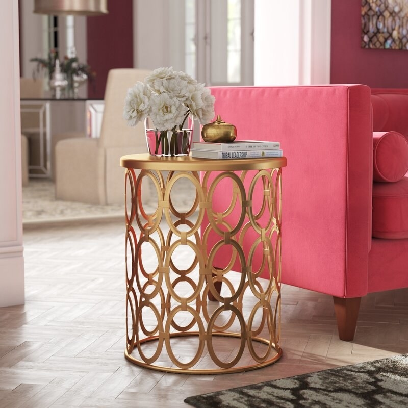 A cylinder-shaped gold end table with metal circles all around the base holding up the top, with books and flowers on top