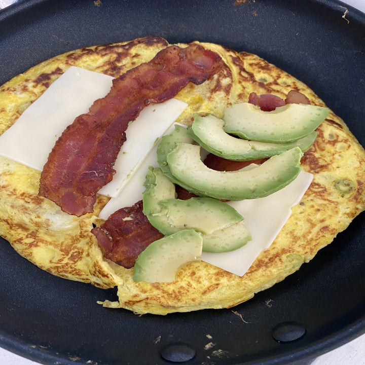 The omelet in a skillet with cheese, bacon, and avocado on top.