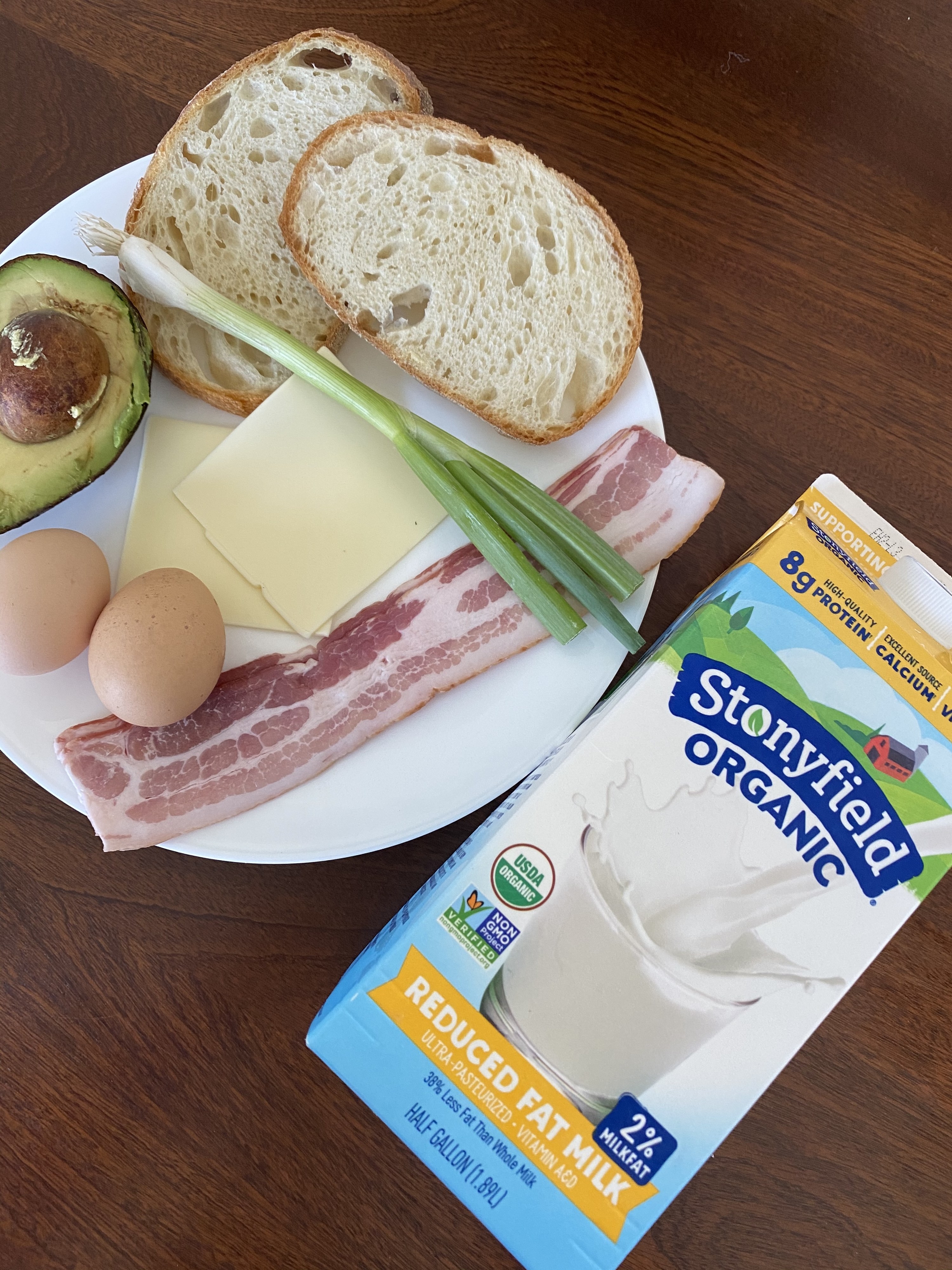 Ingredients for a breakfast sandwich on a countertop, including bread, eggs, bacon, cheese, scallions, avocado, and milk.