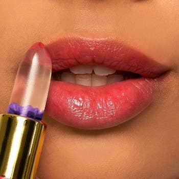 A model applying the balm to their lips, leaving a natural-looking pink tint. The bullet is shaped like a lipstick and is clear and jelly-like, with a purple flower suspended in it