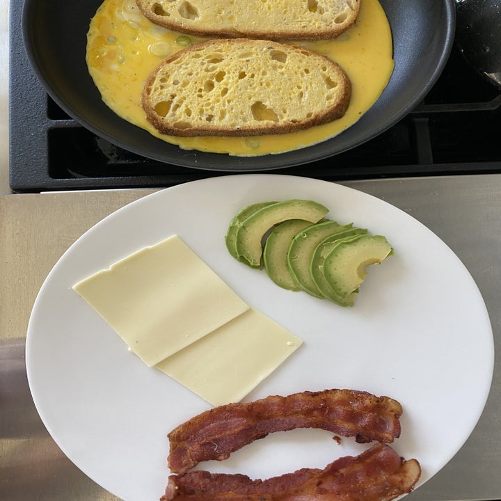 All of the breakfast sandwich ingredients: Bread soaking in scrambled eggs, sliced avocado, cooked bacon, and sliced cheese.