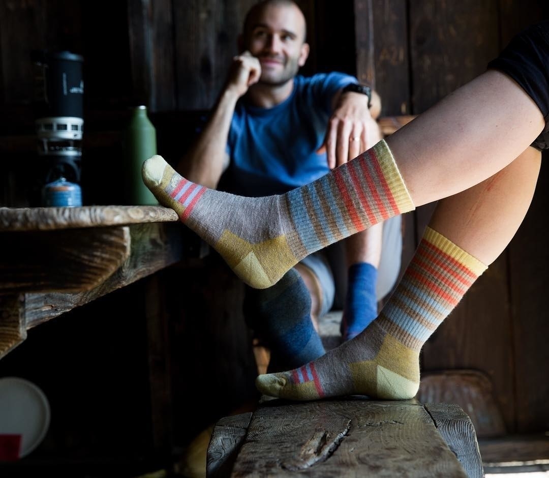 A pair of feet resting on a picnic table wearing the crew-height socks with stripes. Although the exact color is no longer in stock, they&#x27;re grey with blue and orange stripes, and yellow-green on the heels and toes