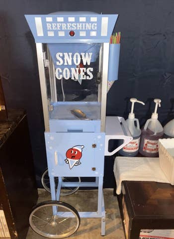 a light blue, tall, vintage-looking snow cone machine with a cute snow cone character on it and two wheels