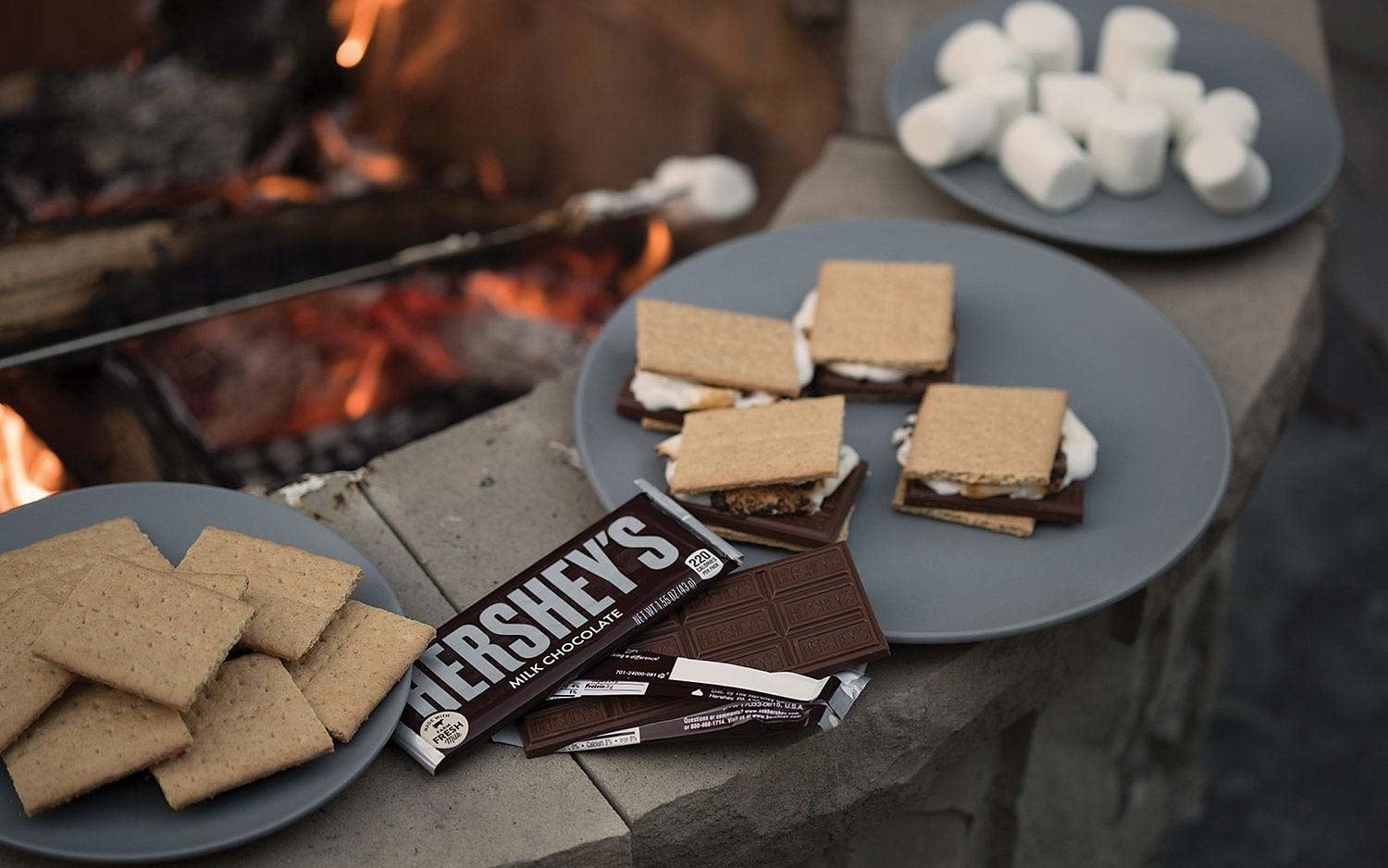 an open hershey&#x27;s chocolate bar next to a plate of s&#x27;mores nearby a fire