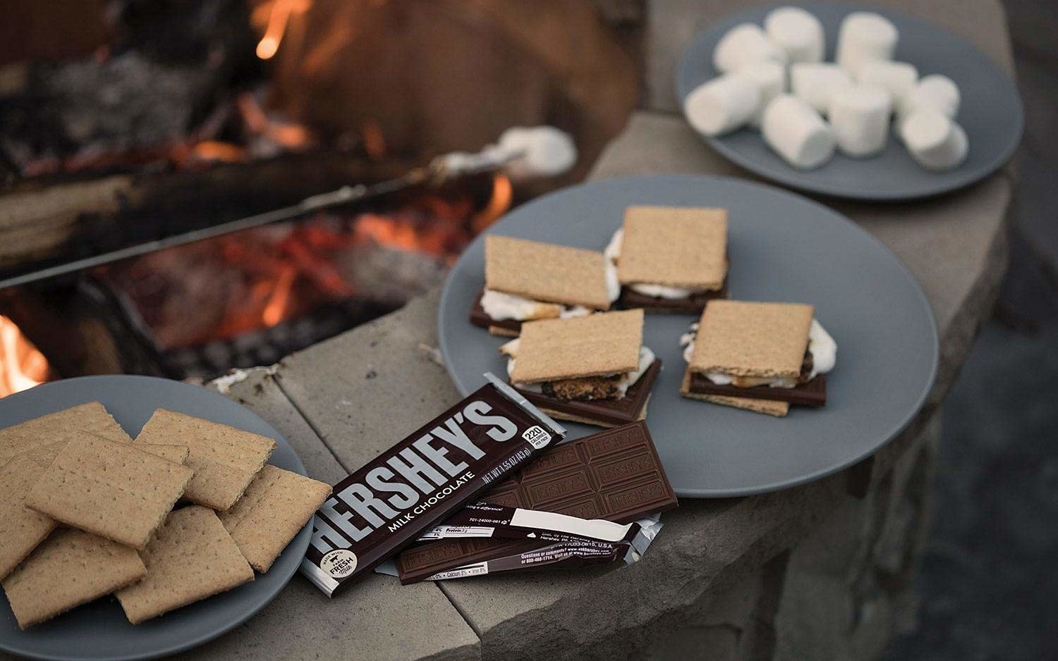 an open hershey's chocolate bar next to a plate of s'mores nearby a fire