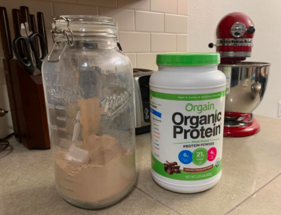 A tall mason jar one third filled with the protein powder sits on a kitchen countertop next to a bottle of Orgain Organic Protein Powder