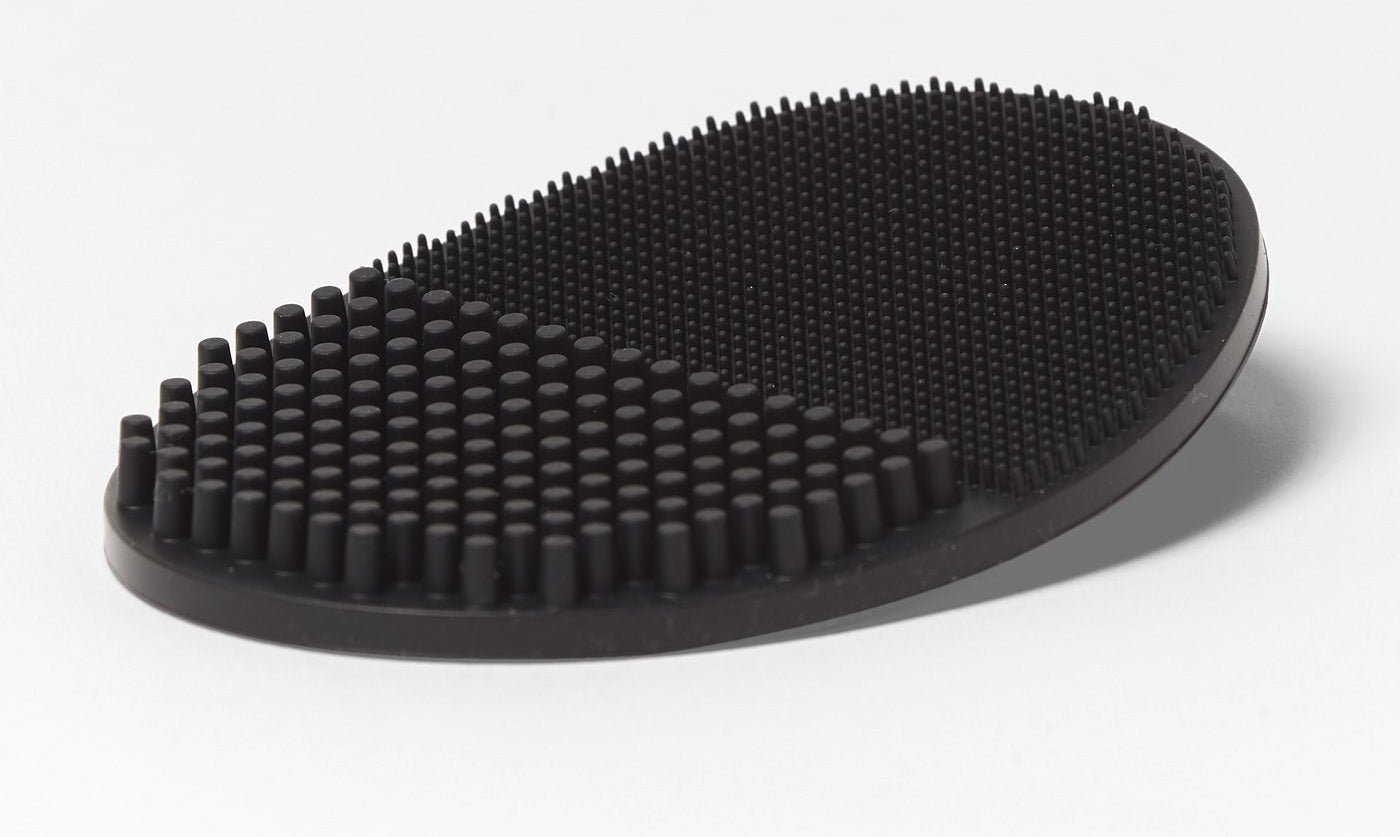 A close-up of a black oval-shaped pad with thick silicone bristles on one side and thinner silicone bristles on the other side