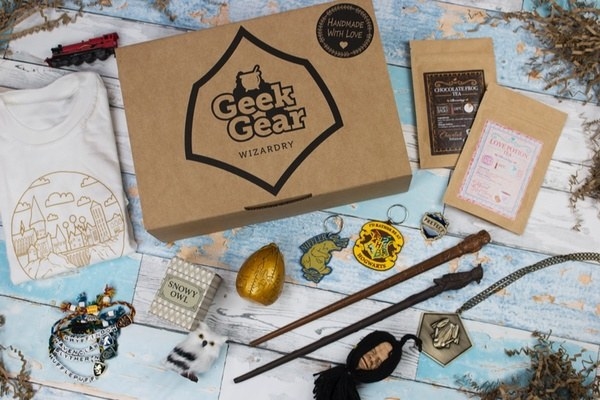 The contents of a box, including two wands, a white folded Hogwarts tee, a golden egg collectible, keychains, bracelets, a necklace, and two postcards