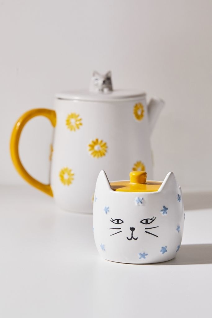 A white ceramic cat-shaped sugar bowl with a delicate blue flower pattern. It has pointy cat ears on top and a bright yellow lid, and it&#x27;s about half the height of the teapot
