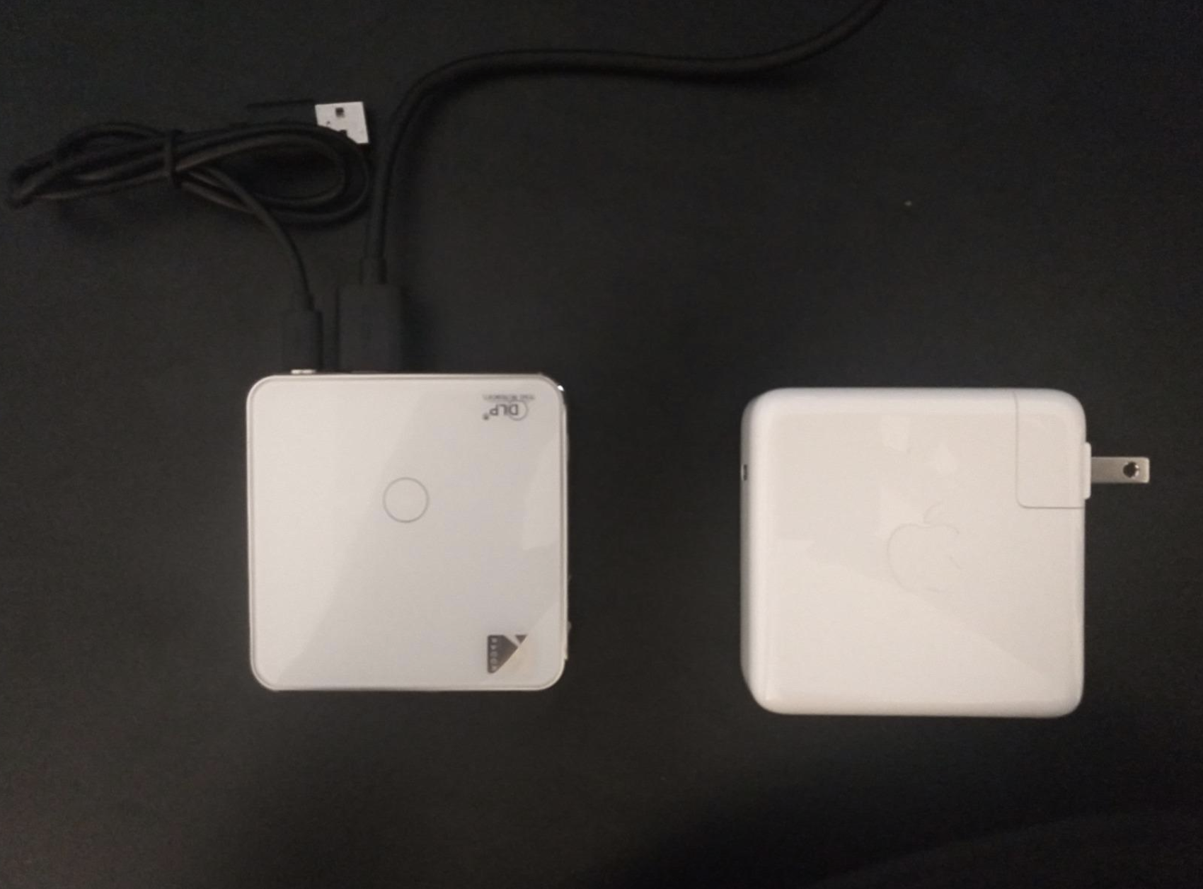 The projector laying on a surface beside a square Macbook charger which is pretty much the same width — no bigger than an average-size coaster