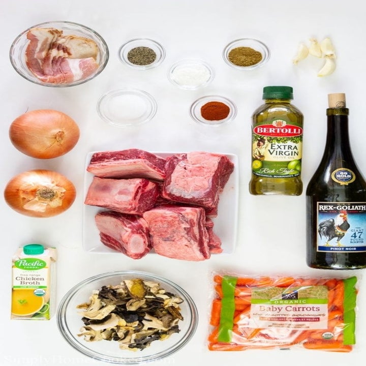 Ingredients for braised short ribs on a countertop, including beef, broth, wine, olive oil, onion, carrots, and spices.