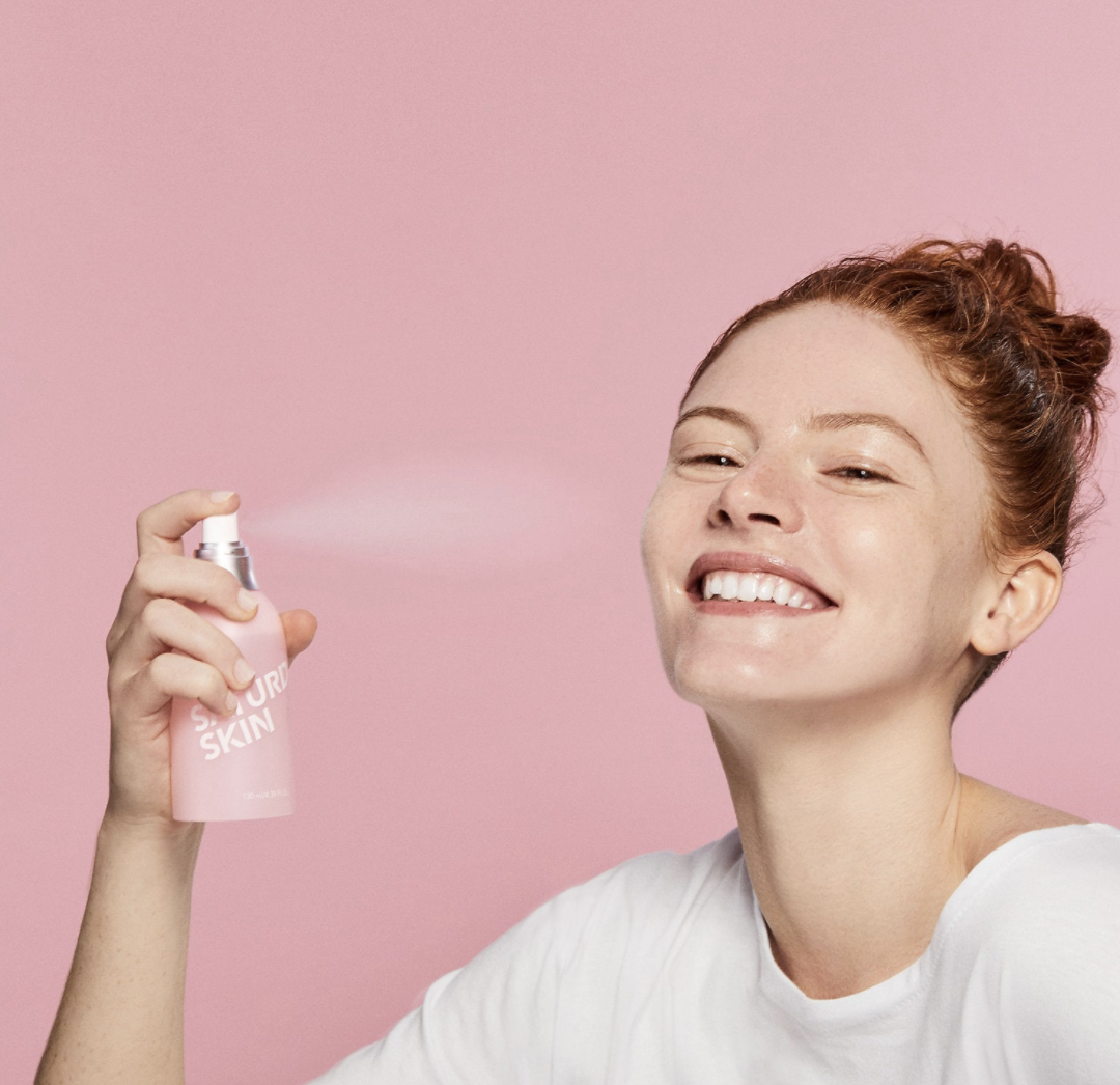 a person holding a pink bottle of saturday skin product and spraying it on their face
