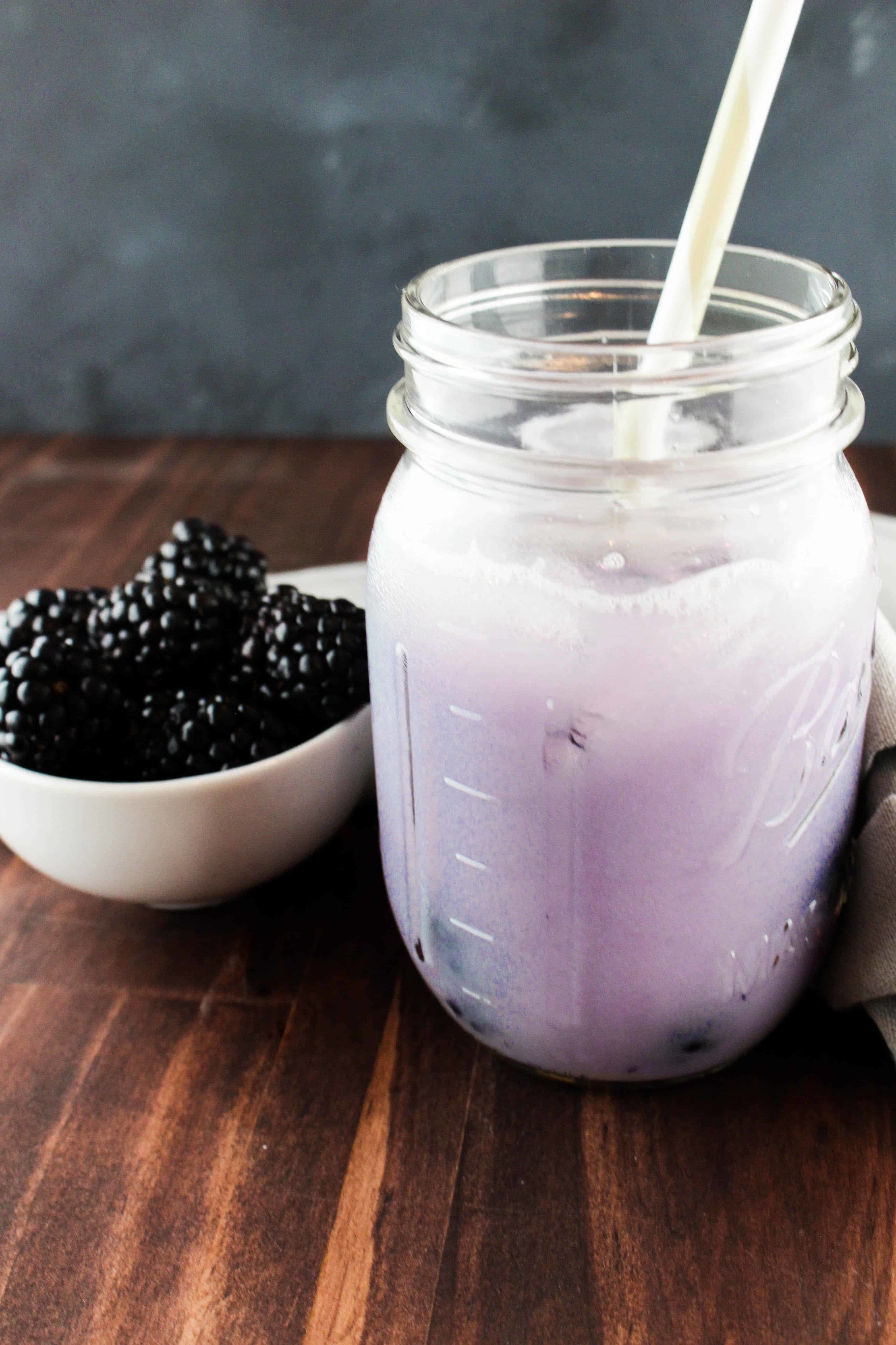 A light purple drink inside a glass Mason jar next to a bowl of blackberries