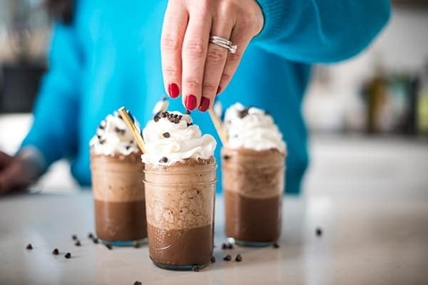 A hand sprinkling mini chocolate chips mason jars filled with the mocha "Frappuccino" and topped with whipped cream