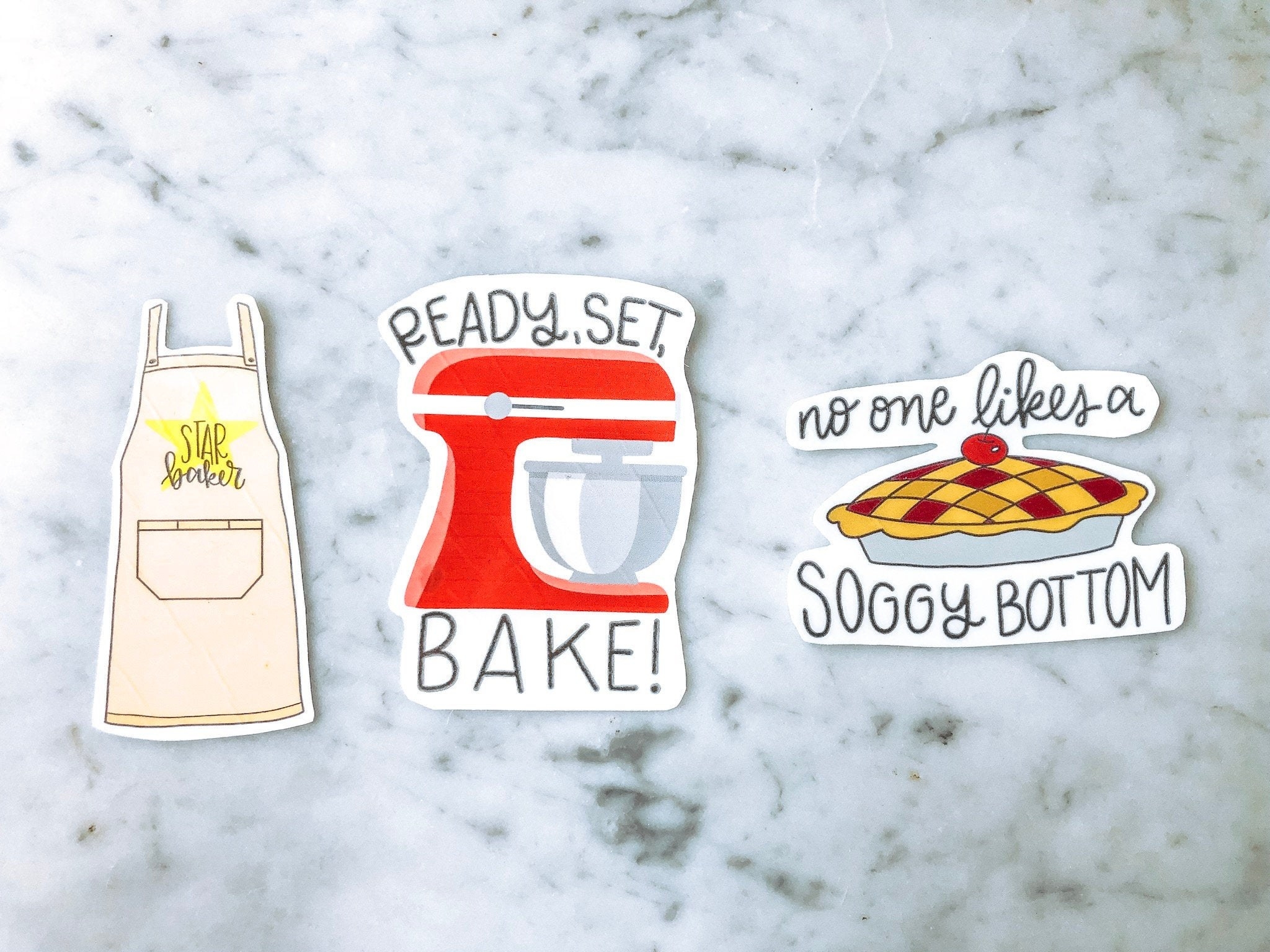 a set of three stickers: the first is an apron that says star baker, the second is a mixer that says ready set bake, the third is a pie that says no one likes a soggy bottom