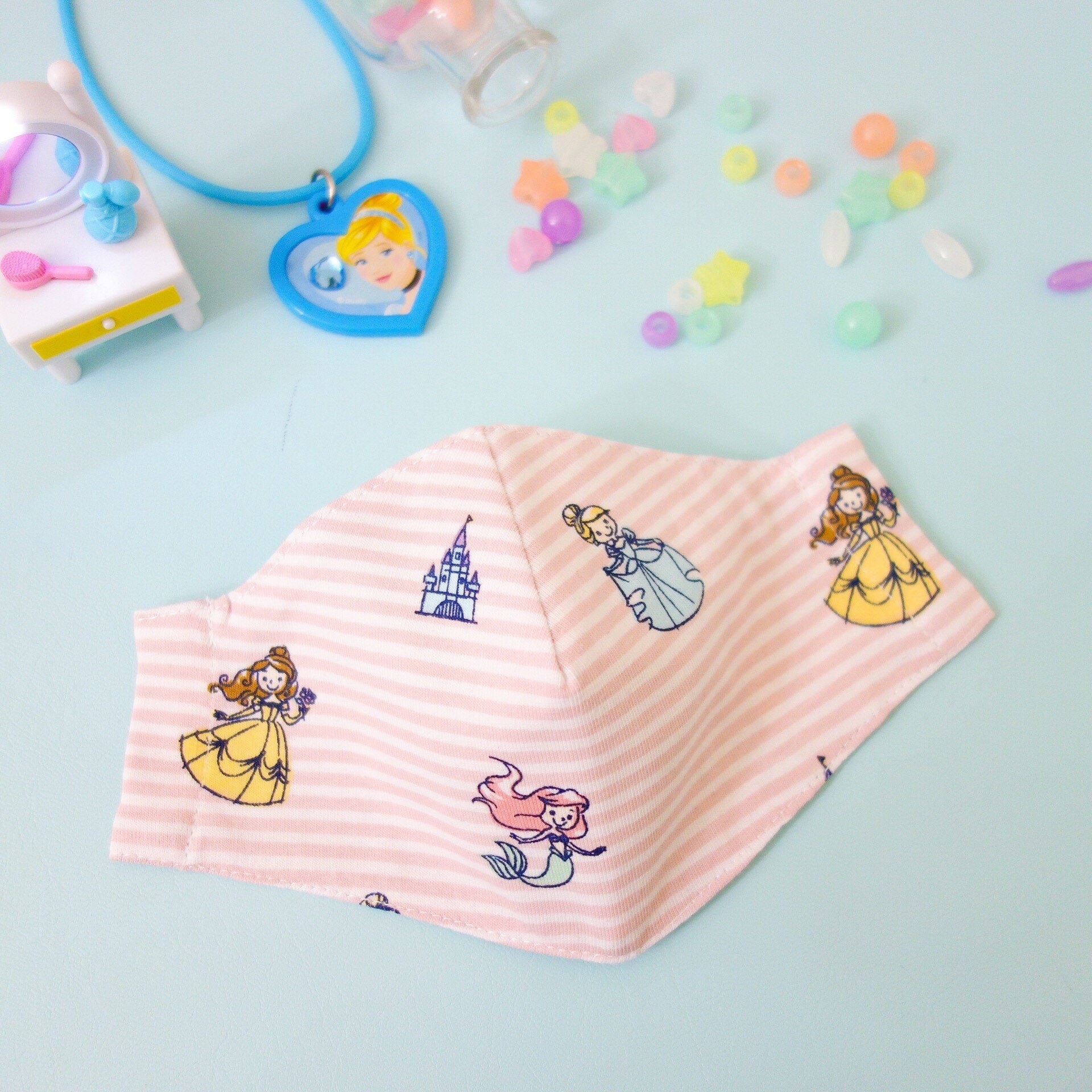 a pastel pink mask with white horizontal stripes and cute illustrations of belle, cinderella, ariel, and the castle
