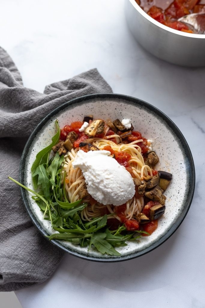 A bowl of spaghetti with eggplant, tomatoes, arugula, and a spoonful of ricotta cheese.
