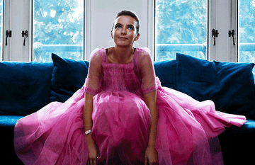Gif of Villanelle from &quot;Killing Eve&quot; In a big organza pink dress sitting on a couch and shrugging her shoulders