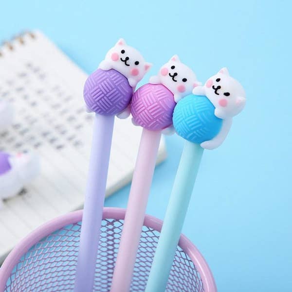 Three of the pens, in lavender, pink, and blue. The top of each pen is shaped like a blushing white cat with a large ball of yarn in the same color as the pen