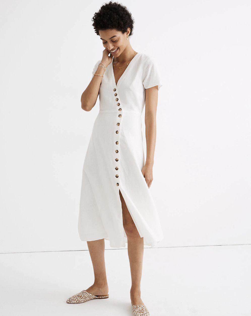 A model in a white linen dress that falls just below the knee, has short sleeves and a v-neck, and buttons all the way up through the v-neck