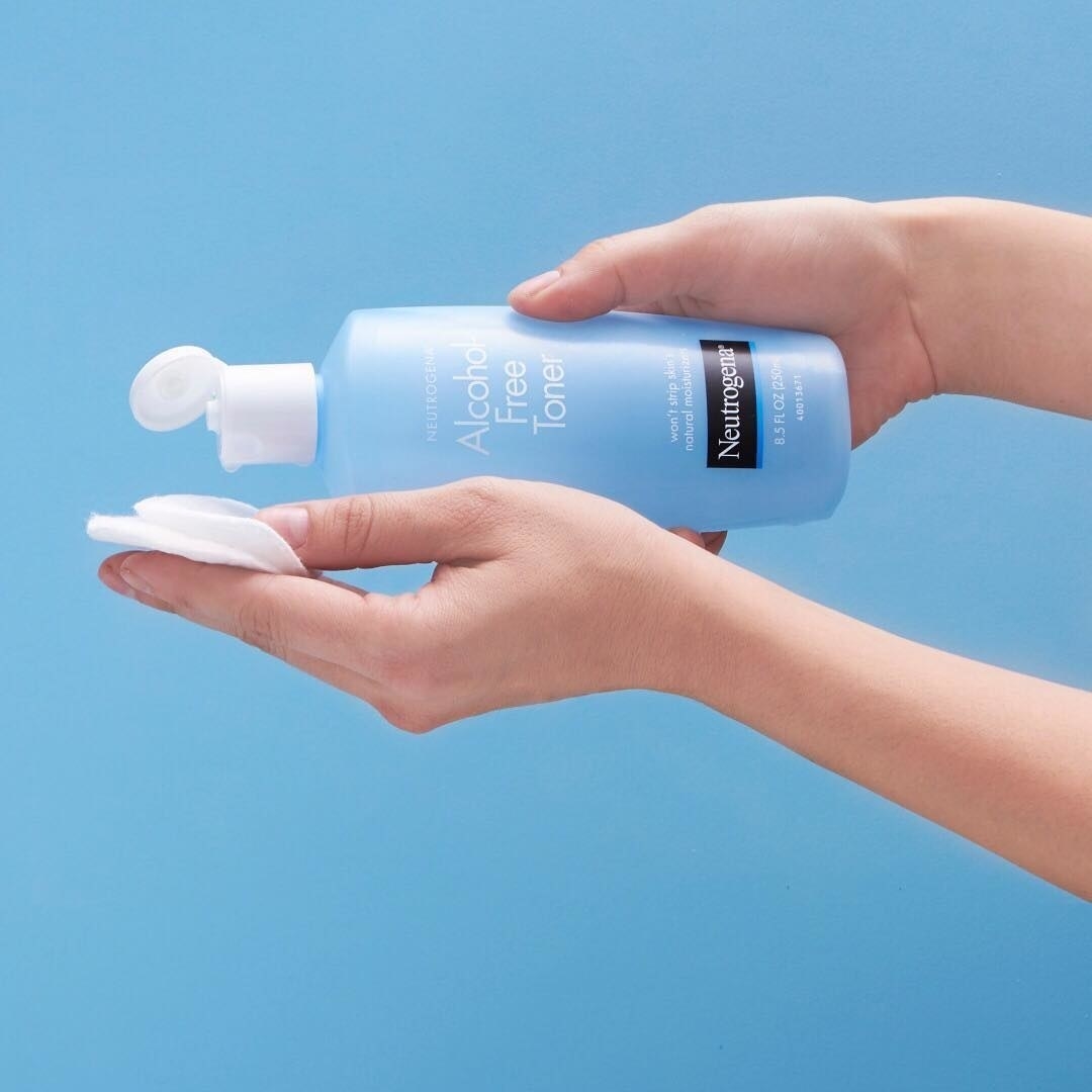 hand holding blue plastic bottle with white cap labeled &quot;Alcohol Free Toner&quot; and &quot;Neutrogena&quot; while the other hand holds out a white cotton round 