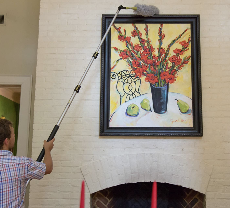 Homeowner uses high-reach dusting kit to wipe away dust from a painting with a vase and fruit over a fireplace