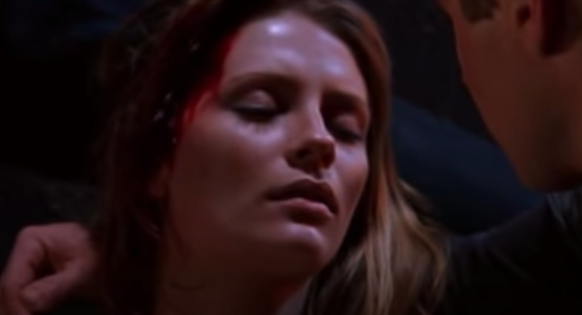 A woman with blood on her head lies on the ground with her eyes shut