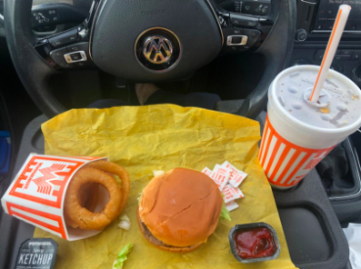 car steering wheel with a tray attached to it containing a fast food meal of a burger, dipping sauce, onion rings, and a drink