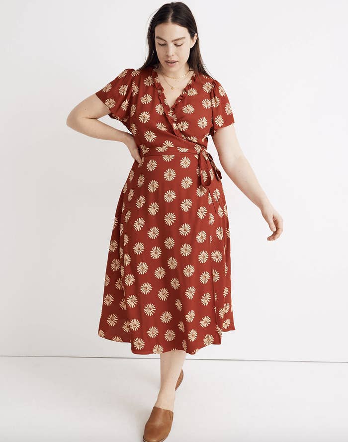 A model in a red dress with daisies that has a V-neck, short sleeves, a tie-string at the waist, and falls mid-shin