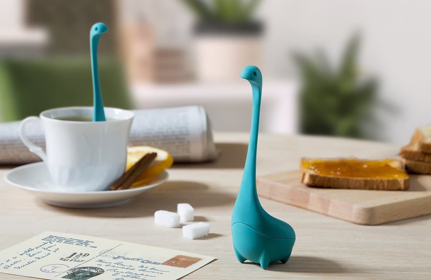 A tea infuser in the shape of a long-neck monster sitting on a table A cup of tea is next to it with another tea infuser inside the cup