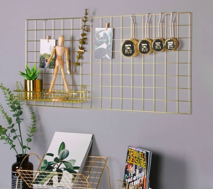 A metal wall hanging with a small attached shelf holding a succulent and doll. There are papers, measuring cups, and flowers clipped onto the grid panel. 