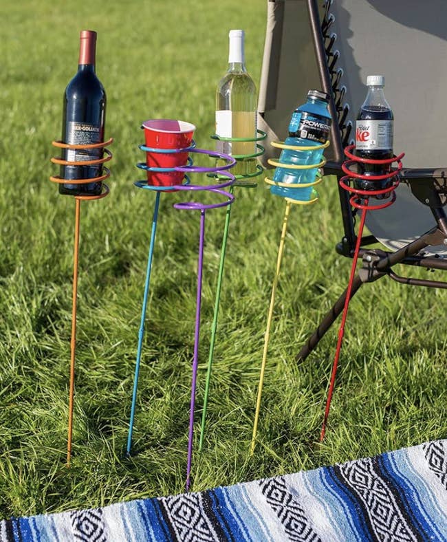 Multi-colored yard drink holders each carrying a bottle of wine, a solo cup, and a Diet Coke bottle next to a woven blanket on the ground