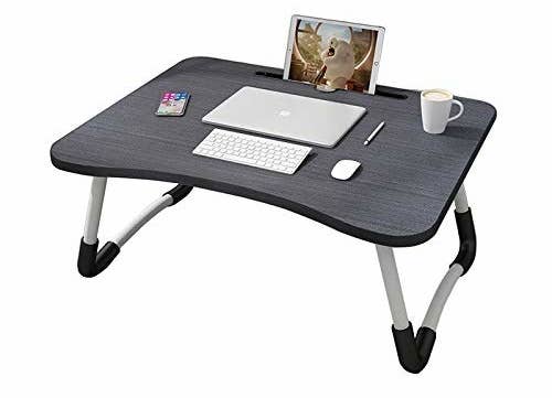 A dark grey foldable bed table with a laptop, wireless keyboard, and mouse on it. Also on the table are a cup of coffee and tablet in the tablet holder slot
