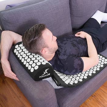 A model lays on the mat and pillow, which is placed on their couch and covered in white circles with small spikes