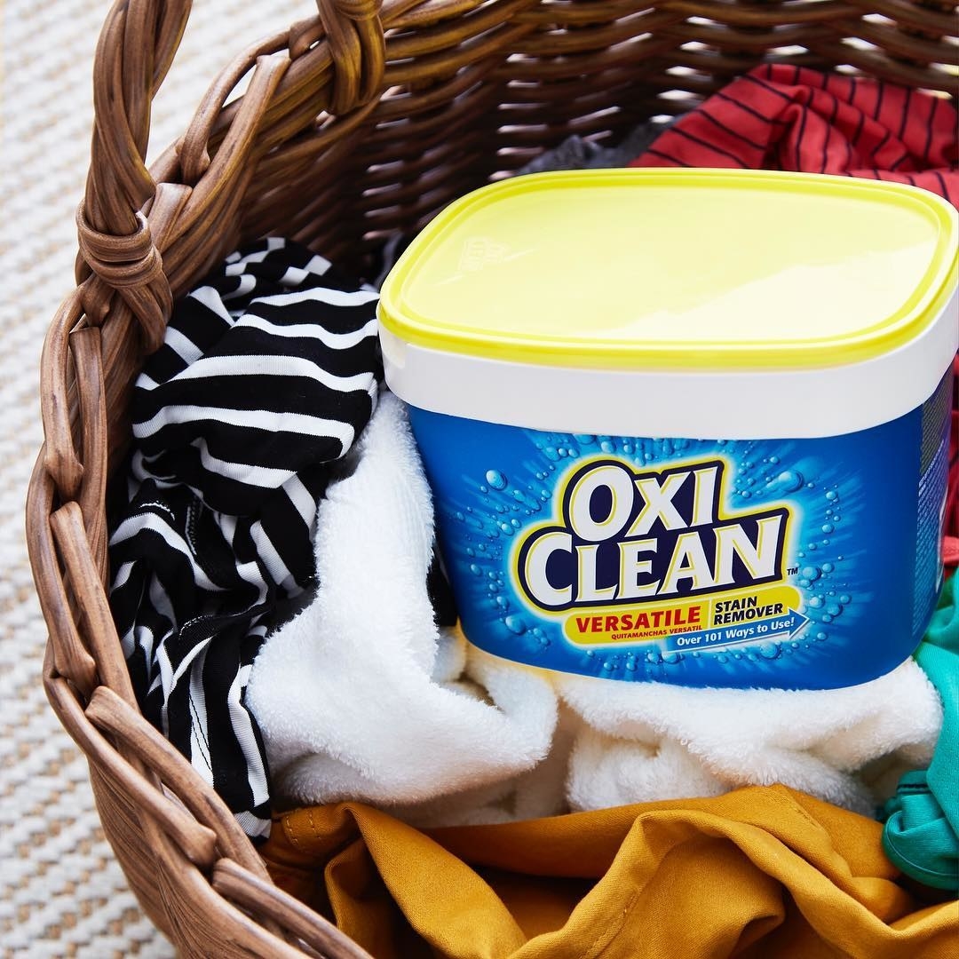 A tub of Oxi Clean stain remover sitting in a laundry basket full of clothes