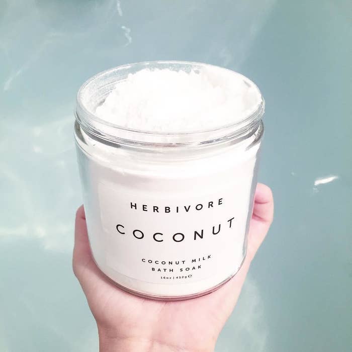 A hand holding a jar of the exfoliant over clear bathwater