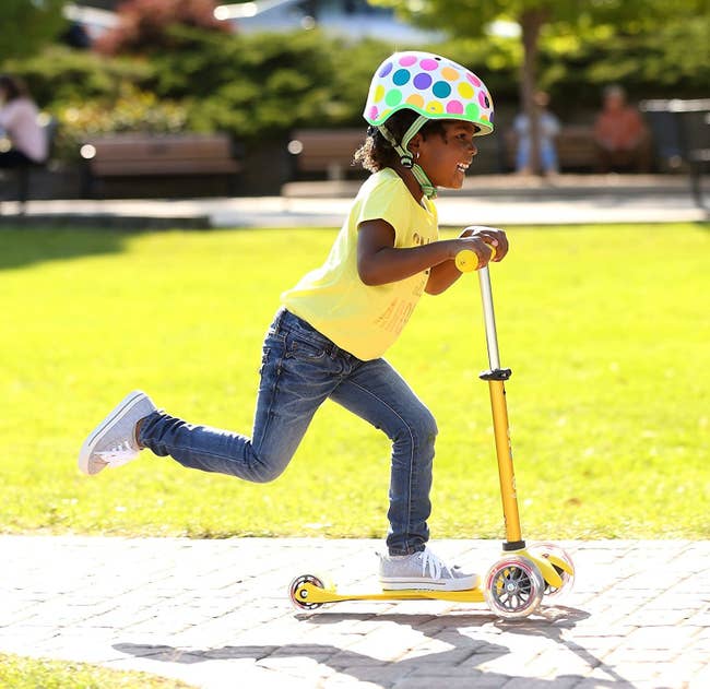 A child model riding a three-wheel scooter outdoors while wearing a helmet
