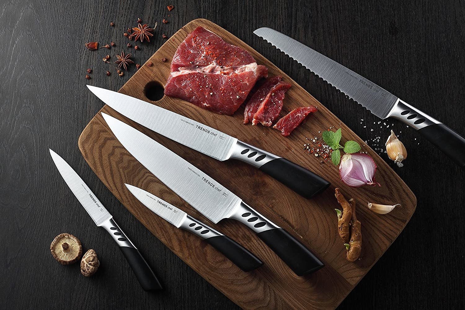 The five knives laying on a cutting board with steak, garlic, mushrooms, and spices