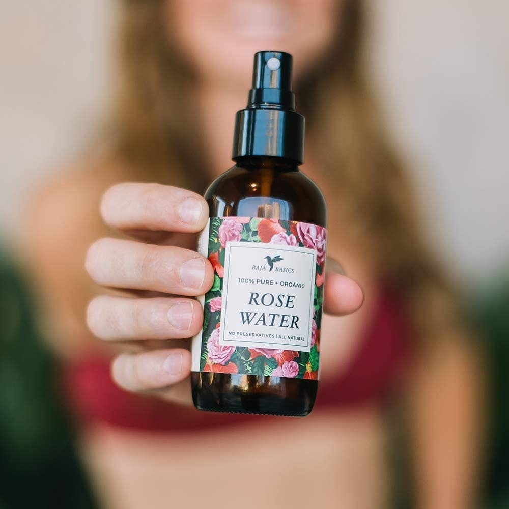 The bottle of rose water toner is seen in sharp relief, held close to the camera by a person who is in the blurry background