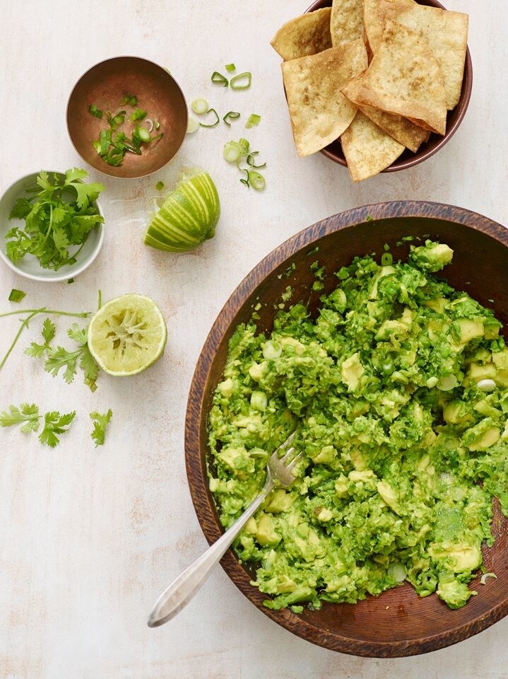 Freshly made guacamole in a wooden bowl with sliced lime, cilantro, scallions, and tortilla chips to the side.