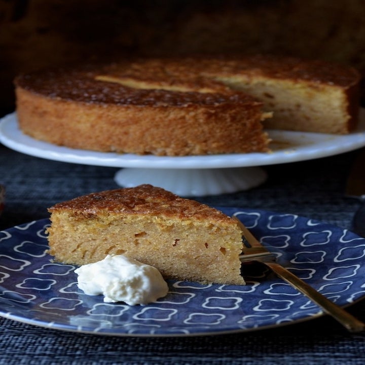 A slice of orange olive oil cake with a dollop of cream on a plate with the full cake in the background.