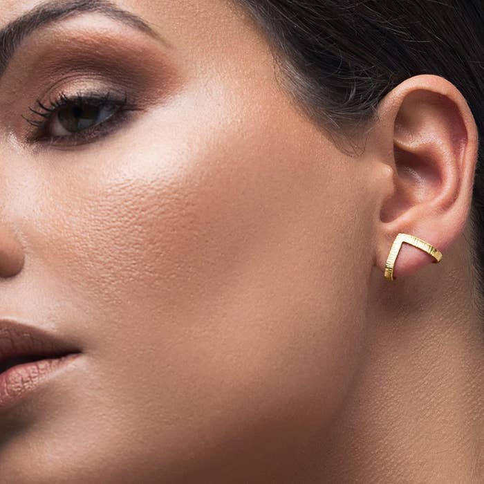 A model wearing one of the stud earrings in the gold finish. It wraps slightly around the back your earlobe and forms a point about where your piercing is, so it&#x27;s a stud that looks like a cuff