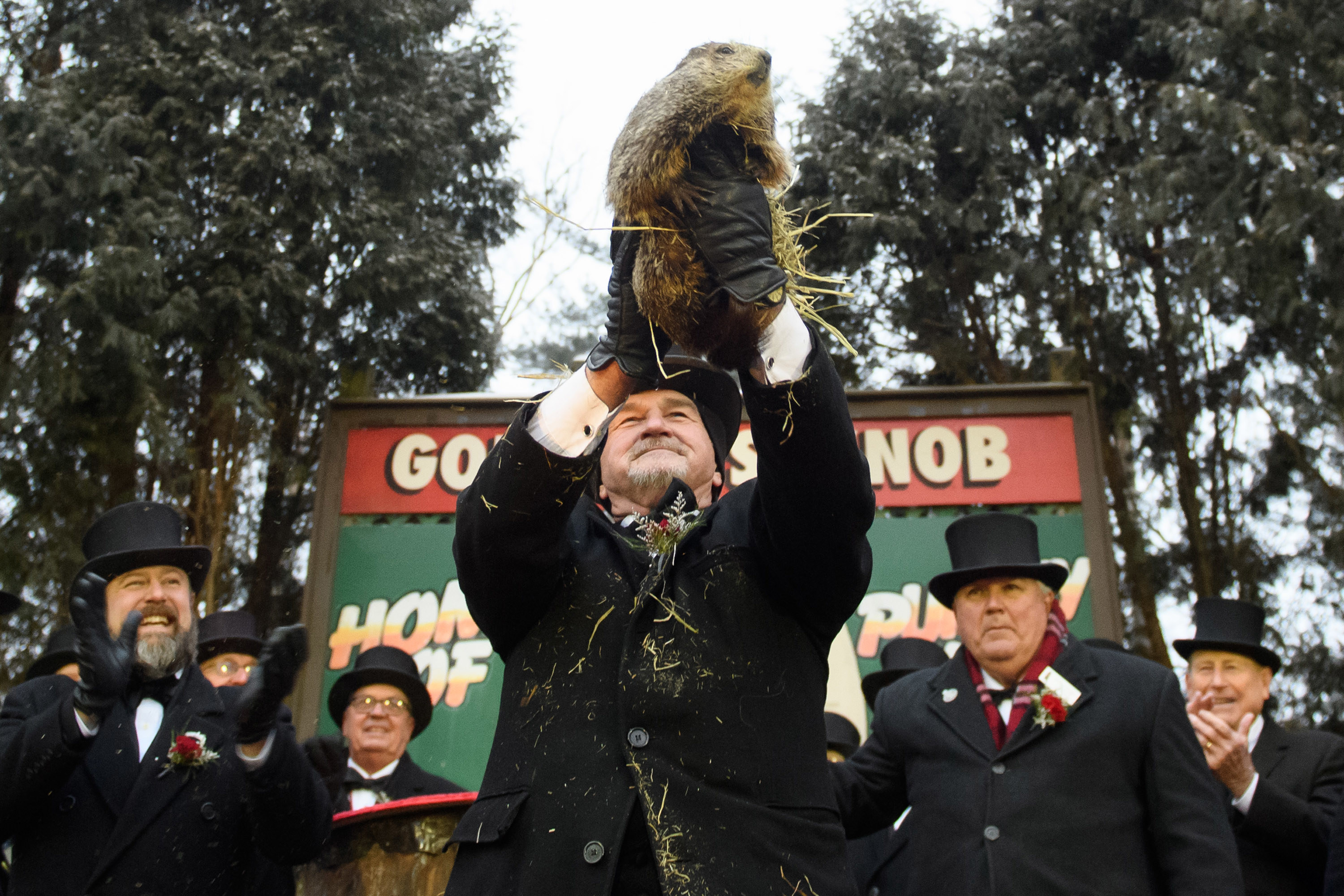 old man holding up a groundhog while men in top hats cheer in the background