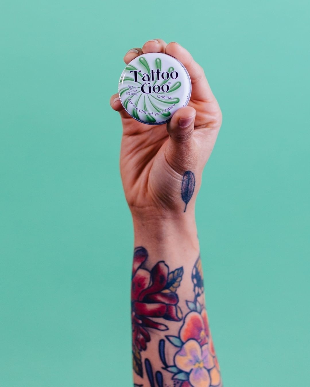 A tattooed hand holds up a tin of the tattoo goo