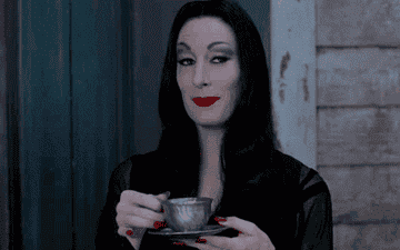 A GIF of Morticia Addams raising a cup of tea and an eyebrow  